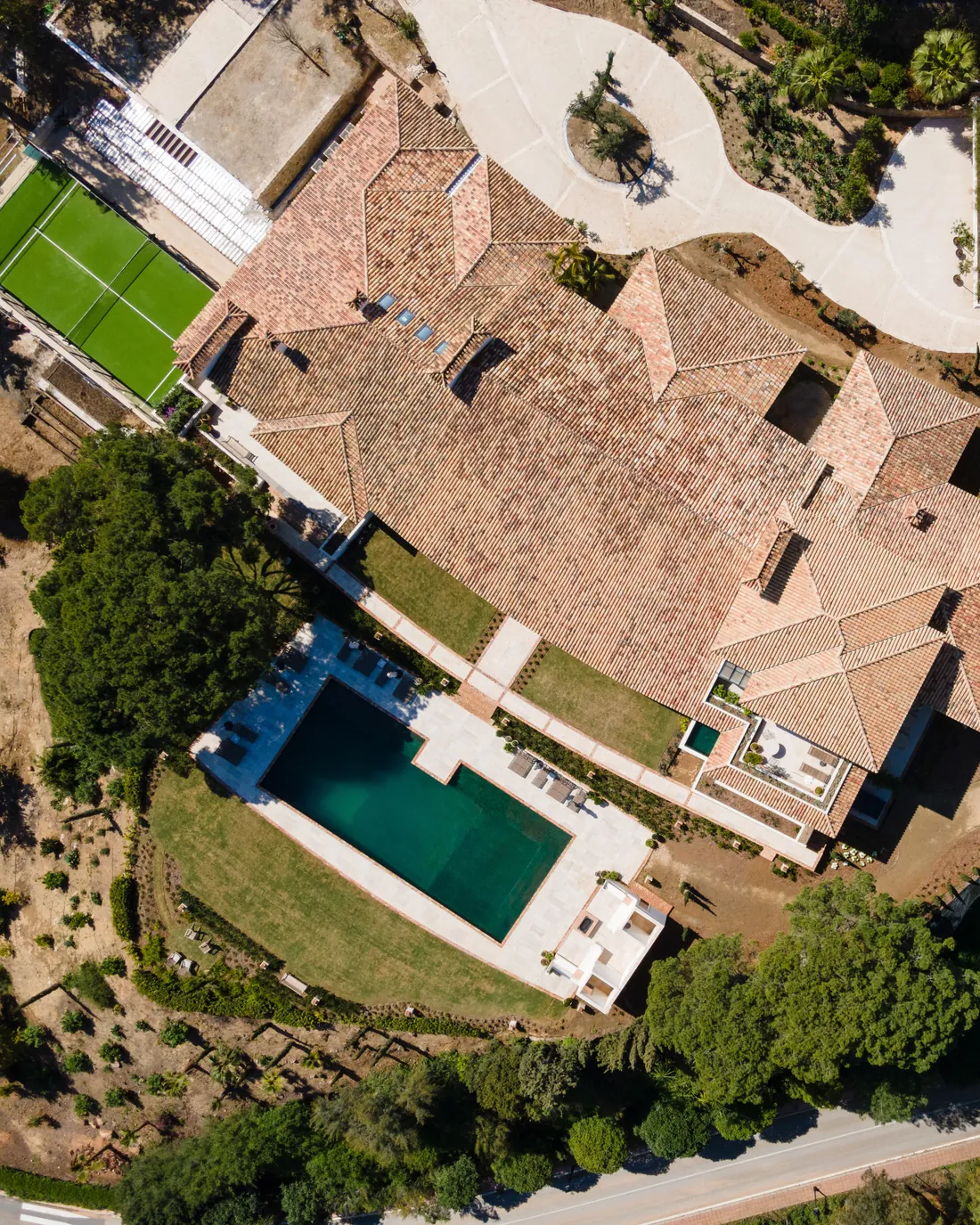 Vista cenital de una villa de cubierta de teja con piscina y pista de pádel en una parcela arbolada